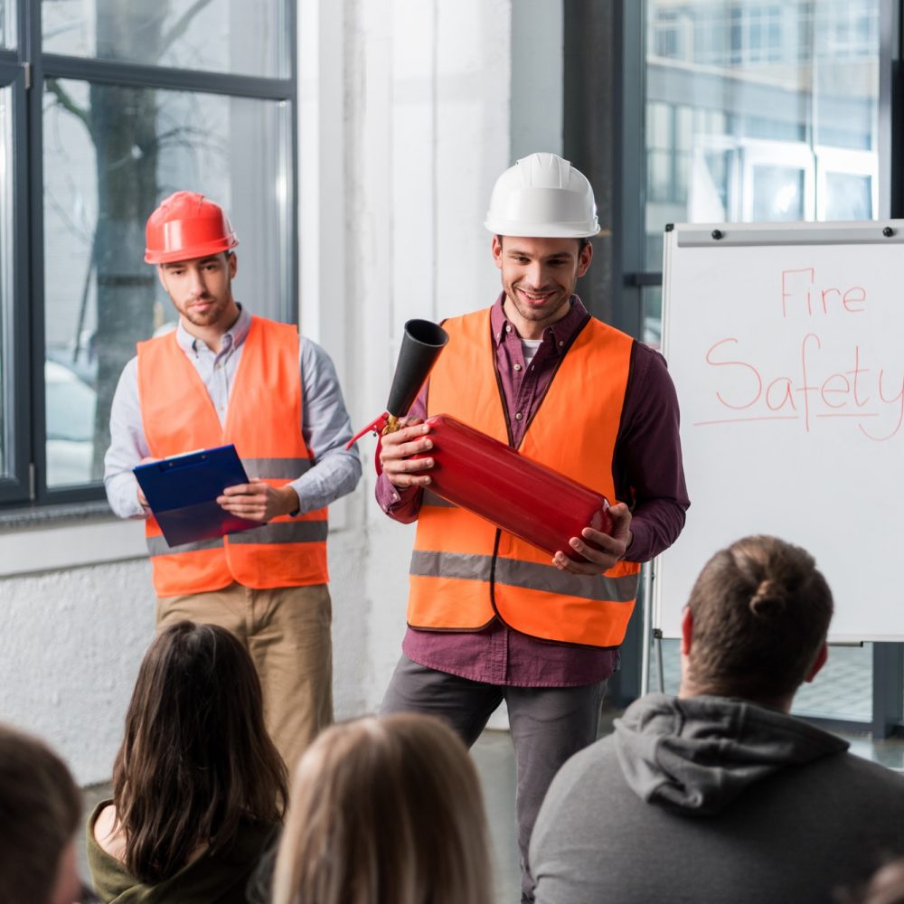 selective focus of handsome firemen in helmets standing near white board with fire safety lettering