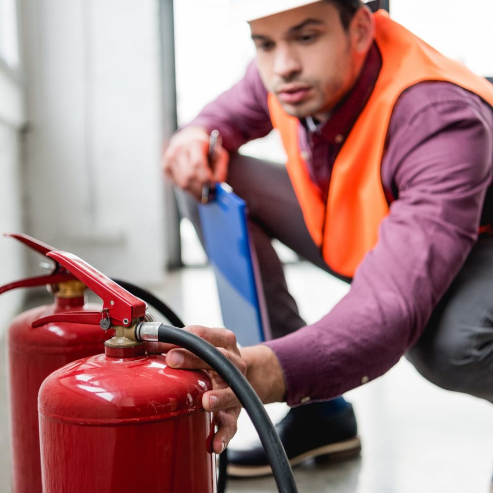 selective focus of red extinguishers near fireman in helmet holding clipboard